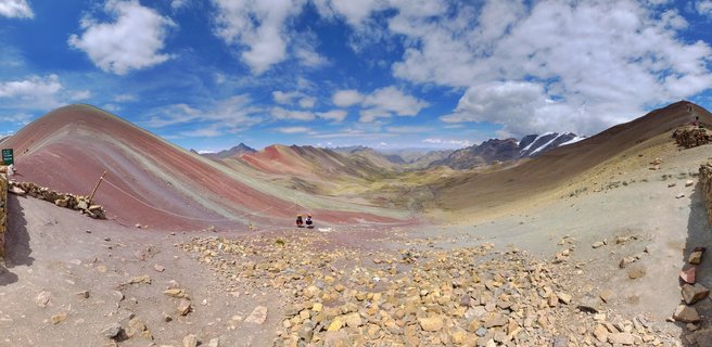 Panoramic view of Rainbow Mountain Vinicunca in Peru showing colorful striped mountain slopes in red pink yellow and green with two small hikers in the rocky foreground under blue sky with white clouds
