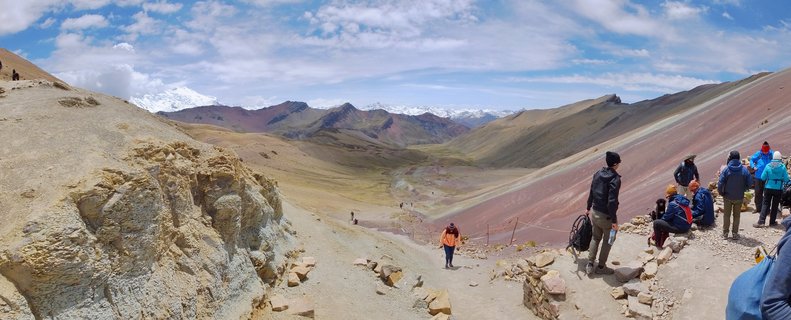 Wide mountain landscape showing tourists hiking on a trail with colorful striped Rainbow Mountain slopes on the right and snow-capped peaks in the distant background under a blue sky with scattered clouds