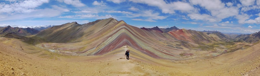Panoramic view of Rainbow Mountain in Peru showing colorful striped mountain slopes with hikers on a dirt trail under blue sky with clouds