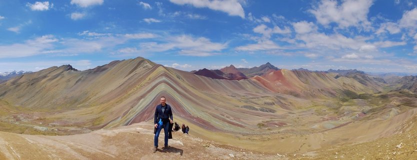 Person standing on mountain ridge with colorful striped Rainbow Mountain (Vinicunca) in background under blue sky with scattered clouds in Peru