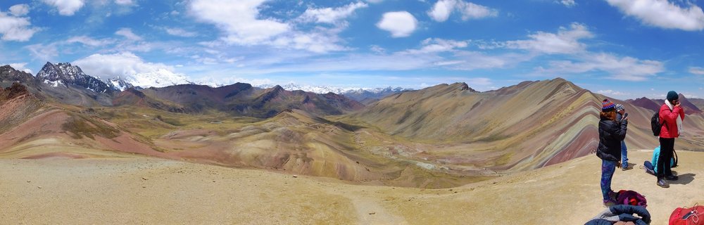 Panoramic view of Rainbow Mountain in Peru showing colorful striped mineral deposits on mountain slopes with tourists on a viewing area and snow-capped peaks in the distance under blue sky with white clouds.