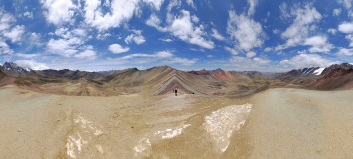 Panoramic view of Rainbow Mountain in Peru showing colorful striped mountain ridges with two hikers in the foreground under a blue sky with white clouds