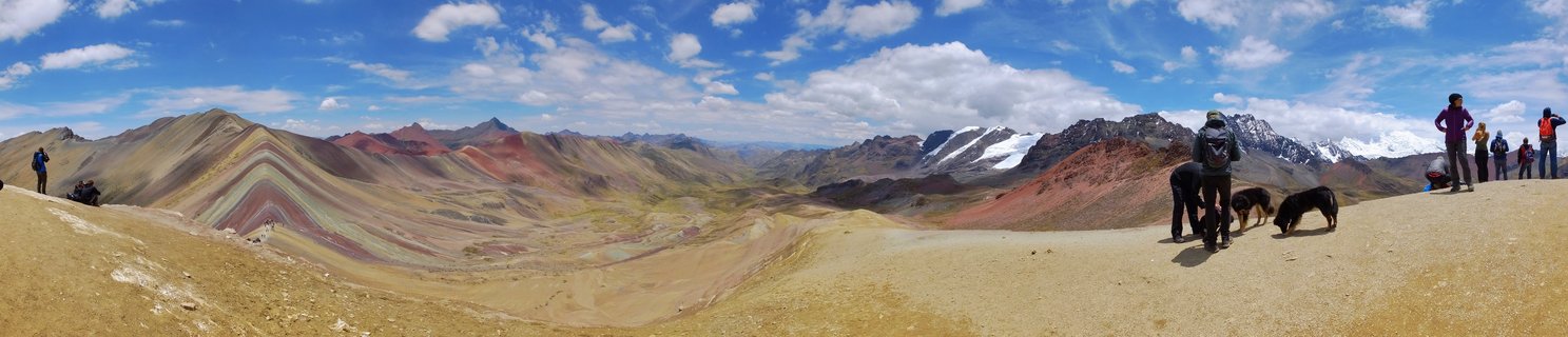 Panoramic view of Rainbow Mountain in Peru showing colorful striped mountain slopes in red, yellow, green, and purple mineral bands, with tourists and llamas at the viewpoint, snow-capped Andes peaks in the distance under blue sky with white clouds