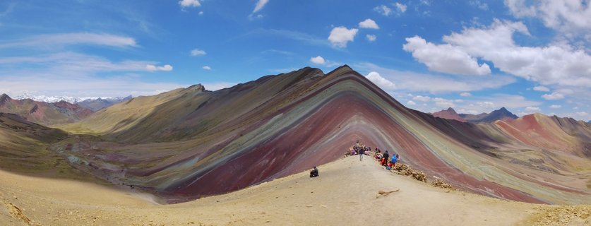 Colorful striped mountain with red, yellow, green and purple mineral layers, tourists on sandy ridge path, snow-capped Andes peaks in distance under blue sky with white clouds
