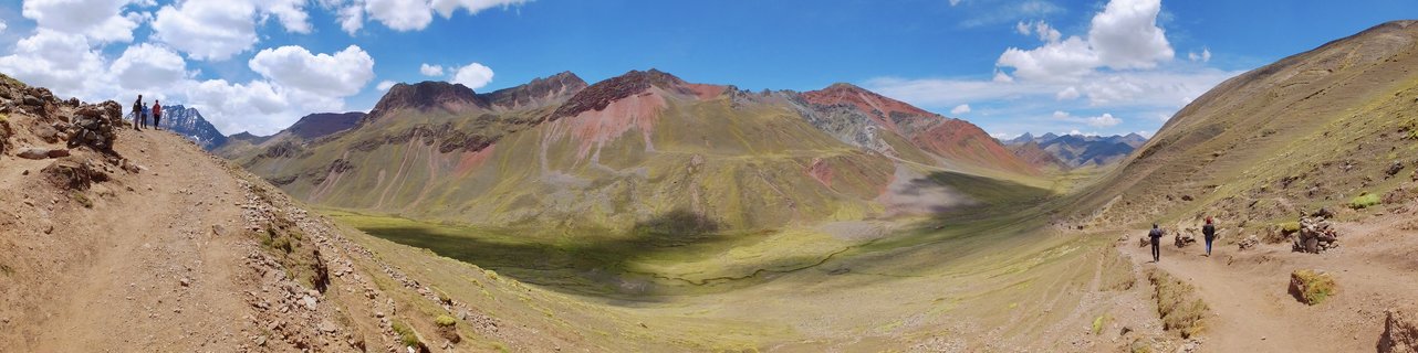 Panoramic landscape showing Rainbow Mountain in Peru with colorful stratified slopes in red, green, and yellow hues, hiking trails with small figures of tourists, green valley below, blue sky with white clouds