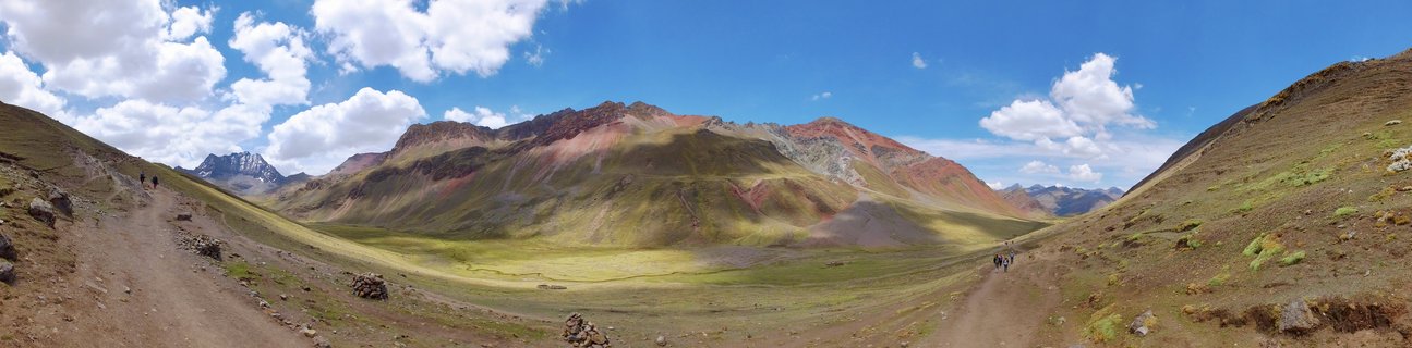 Panoramic view of Rainbow Mountain in Peru showing colorful striped mountain slopes with red, yellow, green and purple mineral layers under blue sky with hikers on dirt trails in valley