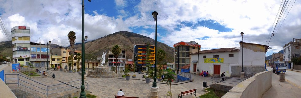 Wide view of a town plaza in Abancay, Peru with a white statue on stone pedestal in center, surrounded by colorful buildings, palm trees, benches with people, and brown Andean mountain in background under cloudy blue sky