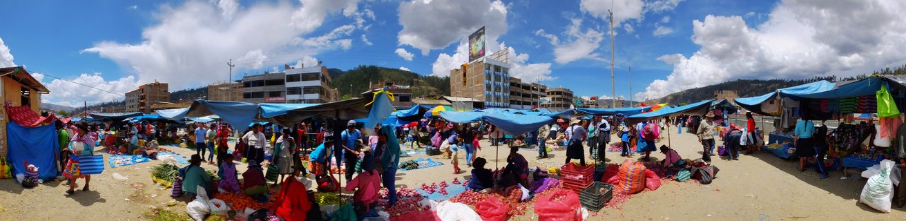 Wide view of an outdoor market in Ayacucho, Peru showing numerous people shopping and selling produce under blue tarps with buildings and mountains in the background
