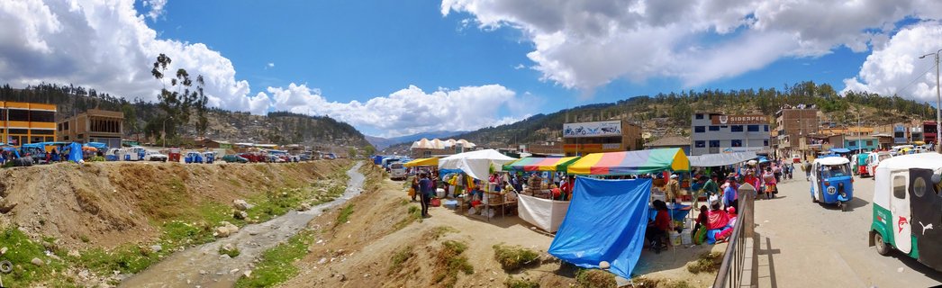 Outdoor market scene in Peru with colorful vendor tents, people shopping, auto-rickshaws on dirt road, small stream, hills and buildings under blue sky with clouds