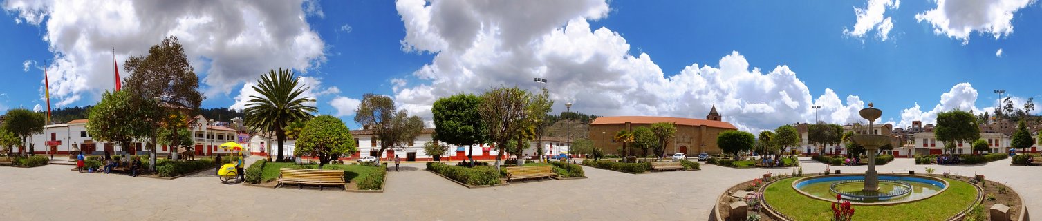 Wide view of a town plaza with a stone fountain, colonial church with red tile roof, white buildings, trees, benches, and people under a blue sky with white clouds