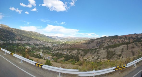 Panoramic landscape showing a valley city surrounded by mountains in Peru, with a white highway guardrail in the foreground and blue sky above