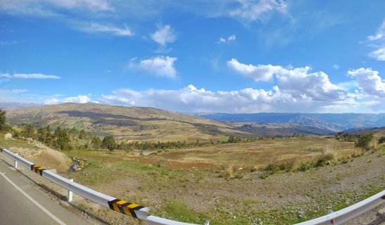 Panoramic landscape showing a paved road with white guardrail through mountainous terrain in Peru's Ayacucho region, with rolling hills and blue sky with white clouds