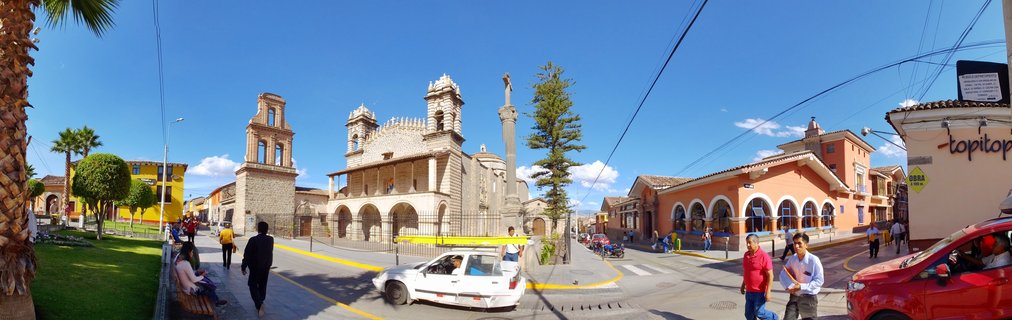 Wide panoramic view of Ayacucho's Plaza de Armas showing colonial cathedral with twin bell towers, central monument, colorful buildings, pedestrians, and vehicles under blue sky
