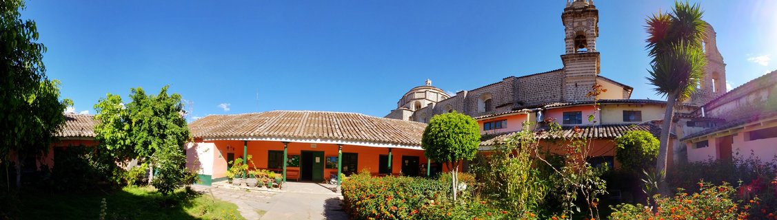 Colonial courtyard with orange and pink buildings, terracotta roofs, green garden, and stone church bell tower under blue sky in Ayacucho, Peru