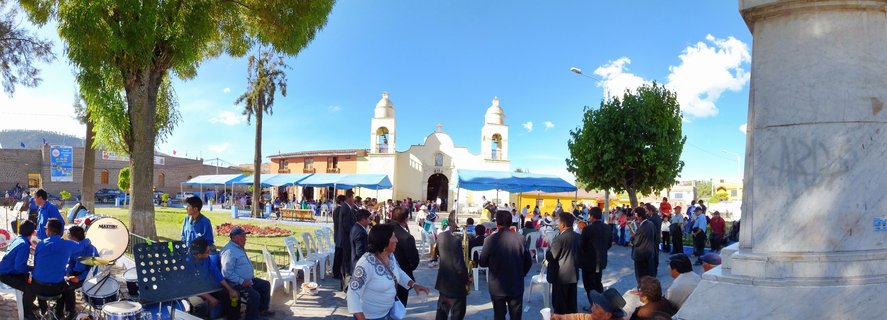 People gathered in a town square in front of a white colonial church with two bell towers, with a traditional band playing drums and blue festival tents set up under a clear blue sky