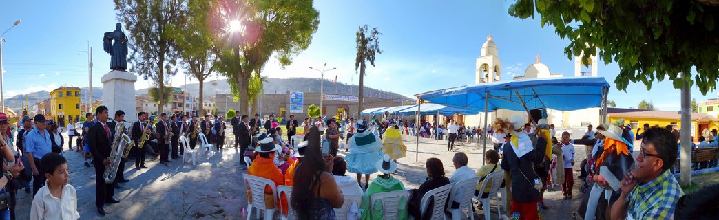 Outdoor cultural festival in a Peruvian town square with brass band musicians, seated spectators, people in traditional Andean clothing, a statue on pedestal, white church with bell towers, and mountains in background