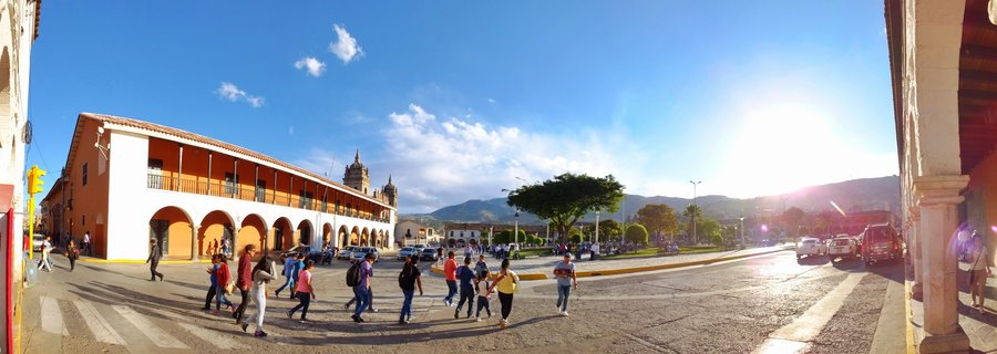 Wide view of a colonial plaza in Ayacucho, Peru showing people walking across a paved square with orange and white arcaded buildings, a baroque cathedral with twin towers, green trees, and mountains in the background under a bright blue sky with afternoon sunlight