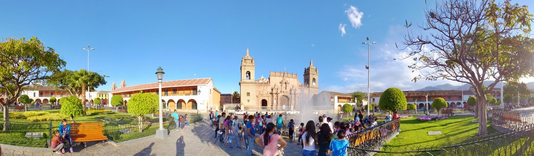 Wide view of a colonial town square with a baroque cathedral, people walking, fountains, gardens, and Spanish colonial buildings with arcades