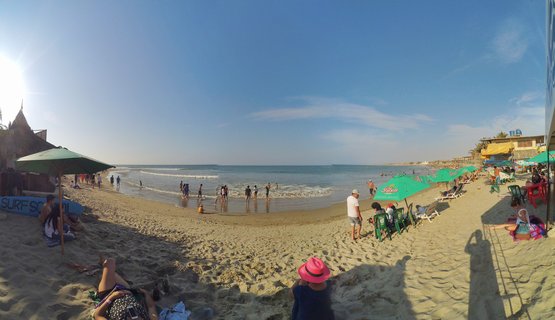 Wide beach scene showing people under green umbrellas on sand with ocean waves and blue sky in Mancora, Peru