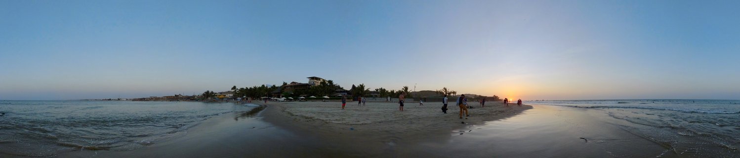 Wide beach panorama showing people walking on a sandbar at sunset with ocean on both sides, palm trees and buildings in the background under a clear gradient sky