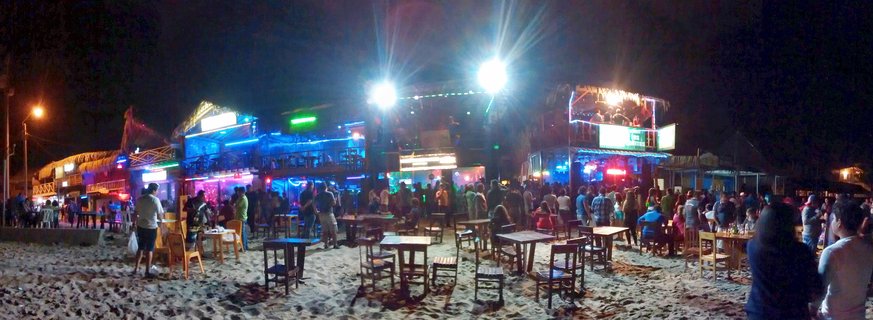 Night panorama of beach bars in Mancora Peru with people sitting at wooden tables on sand under bright colorful lights and neon signs