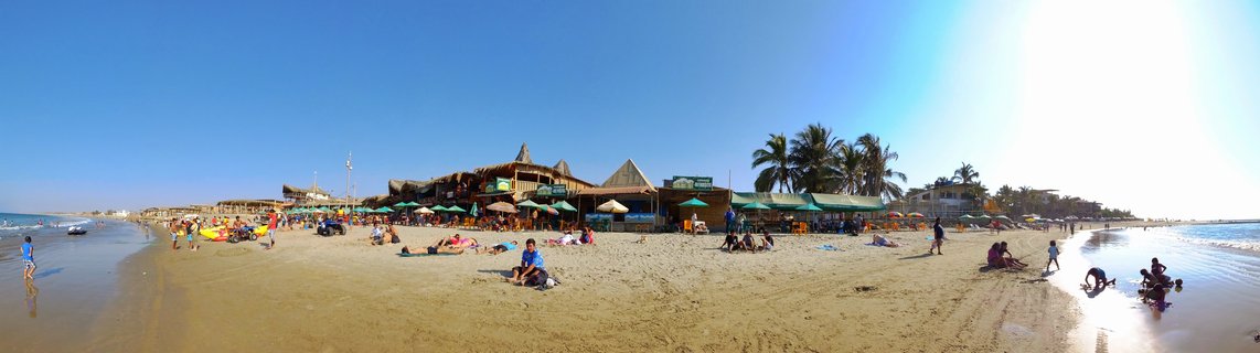 Wide panoramic beach scene showing Mancora Peru coastline with people on sand, beach restaurants with thatched roofs, green umbrellas, palm trees and blue ocean under clear sky