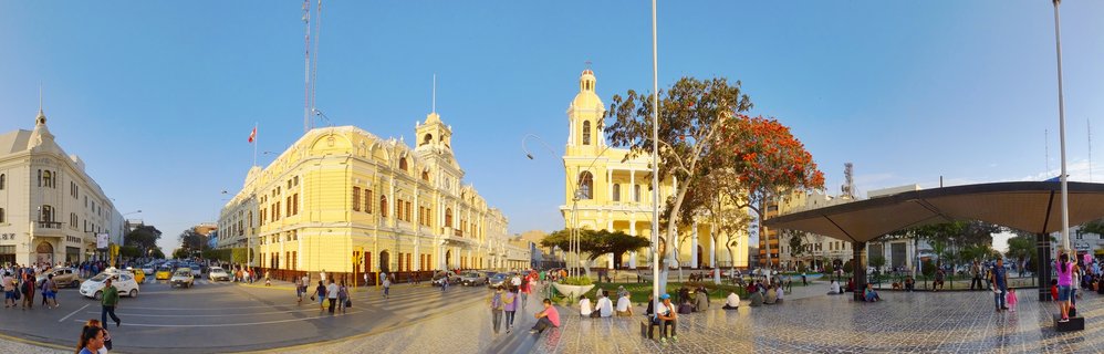 Panoramic view of a city plaza in Chiclayo, Peru with yellow colonial buildings, a church bell tower, people walking and sitting, vehicles on the street, and clear blue sky