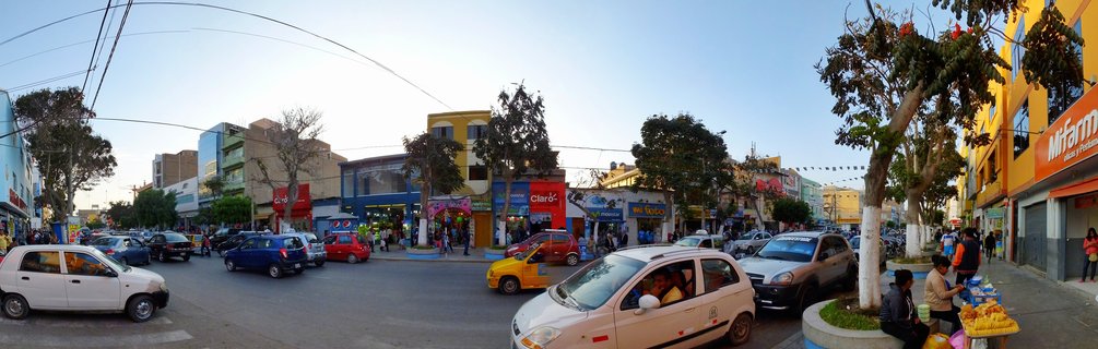 Busy street intersection in Chiclayo, Peru showing colorful buildings, multiple cars and taxis, street vendors with fruit stands, trees along sidewalks, and pedestrians during late afternoon