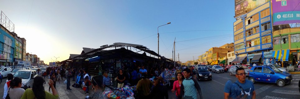 Wide street view of an outdoor market in Chiclayo, Peru with covered vendor stalls, pedestrians shopping, parked cars, and colorful multi-story buildings under a clear blue sky