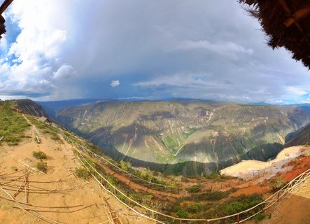 Wide canyon landscape view from elevated viewpoint in Chachapoyas Peru showing green mountain slopes with cloud shadows dirt path with wooden railings in foreground and dramatic cloudy sky