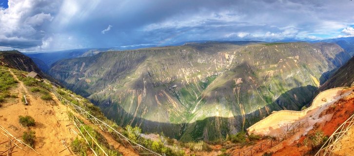 Panoramic canyon landscape with steep green and brown mountain walls, wooden railings along a dirt viewpoint path, dramatic clouds with sun rays breaking through, Chachapoyas region Peru