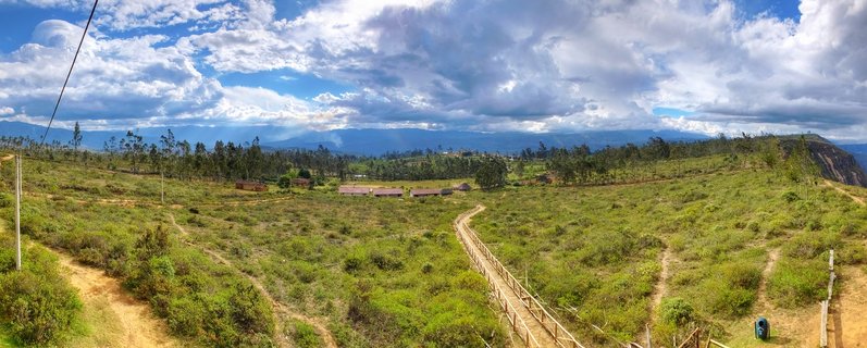 Wide landscape view of green highland valley in Chachapoyas Peru with wooden boardwalk pathway, small buildings with red roofs, scattered trees, distant blue mountains, and cloudy sky