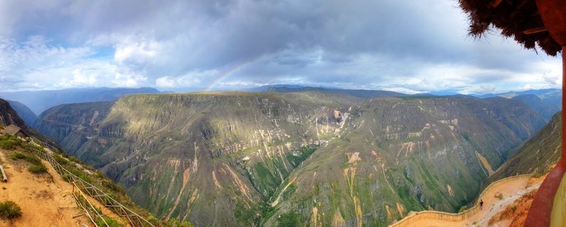 Panoramic landscape showing a deep green canyon valley in Peru with steep mountain slopes, dramatic cloudy sky with a rainbow, hiking trails with wooden railings in foreground, and small thatched-roof structures
