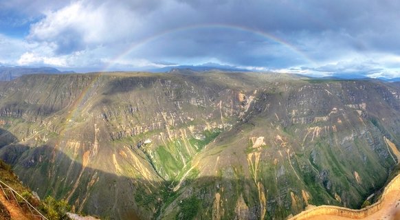 Panoramic view of a mountain canyon with a rainbow arching across cloudy sky over green and brown valley slopes in Peru