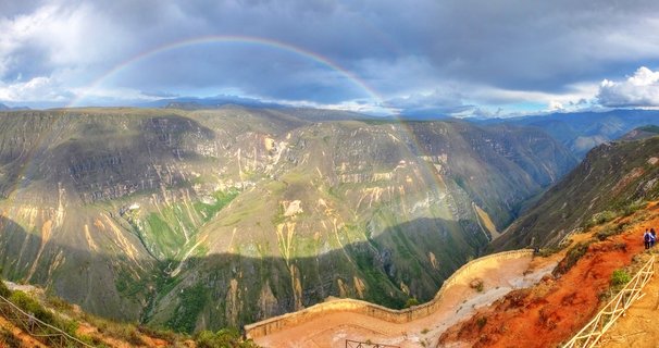 Panoramic view of a rainbow arching over a deep mountain canyon with steep green and rocky slopes, dramatic cloudy sky, and a viewpoint with stone walls in the foreground near Chachapoyas, Peru