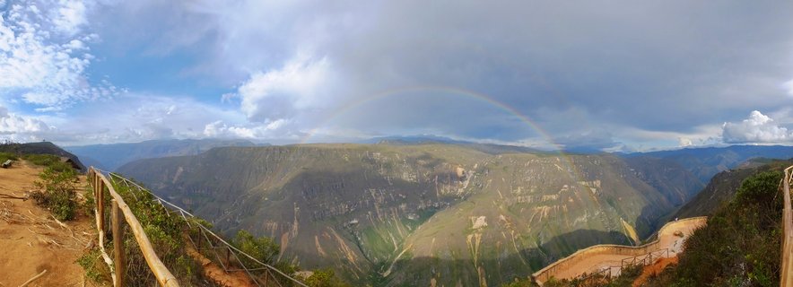 Panoramic view of a deep mountain canyon in Peru with a double rainbow spanning the cloudy sky, wooden fence railings visible at the viewpoint in the foreground