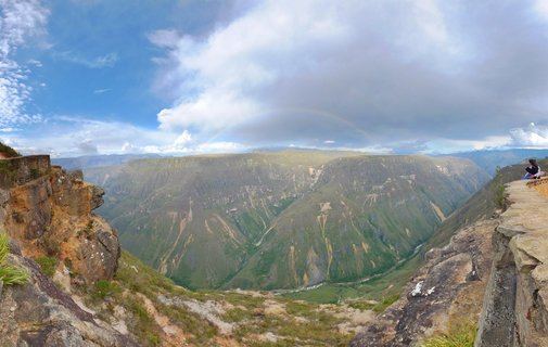 Panoramic view of a deep green canyon valley in Peru with a faint rainbow in the cloudy sky above, rocky cliff edges in the foreground, and a person sitting on the right cliff overlooking the landscape