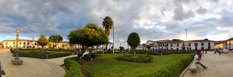 Wide view of a town plaza with green lawns, trimmed hedges, trees, people on benches, white colonial buildings, and a twin-towered church under cloudy sky