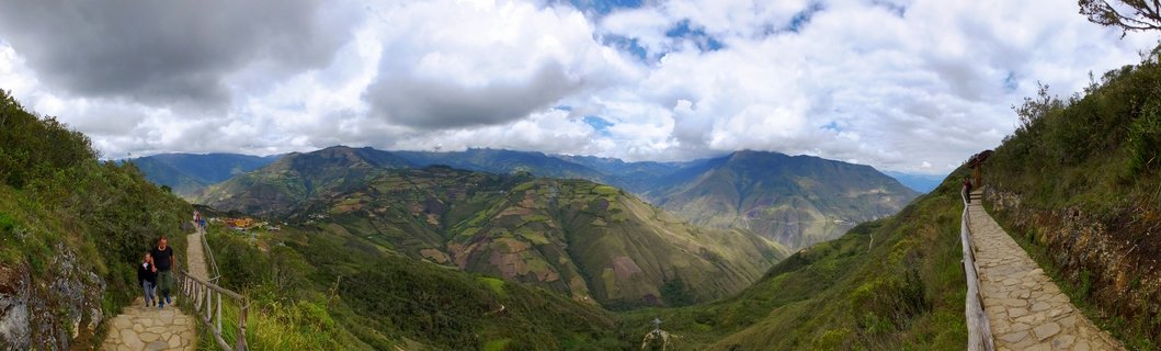 Panoramic view of Kuelap archaeological site showing stone pathways with wooden railings where tourists walk, surrounded by lush green mountainous terrain with agricultural terraces, deep valley, and dramatic cloudy sky