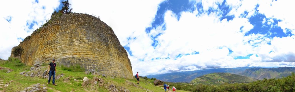 Large curved ancient stone wall structure on green hillside with several people walking nearby, blue sky with white clouds above, mountains visible in background distance