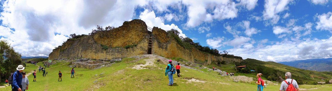 Ancient stone fortress walls of Kuelap archaeological site with tourists walking on green grass under blue sky with white clouds