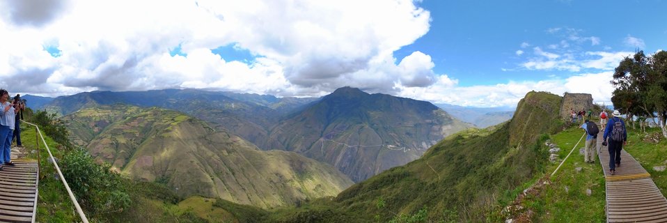 Panoramic view of Kuelap ancient fortress in Peru showing stone ruins on mountain ridge with tourists walking on wooden boardwalks, surrounded by green Andean valleys under blue sky with white clouds