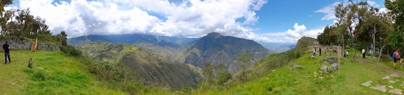 Wide panoramic landscape showing ancient stone fortress walls on grassy hilltop with tourists walking on paths, overlooking deep green mountain valley under blue sky with white clouds in Kuelap, Peru