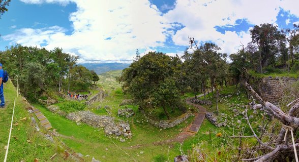 Aerial view of ancient stone ruins at Kuelap archaeological site with green grass, trees, wooden walkways, and visitors exploring the Chachapoya fortress in Peru