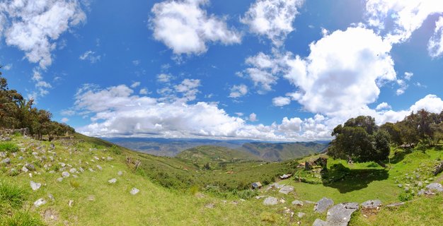 Panoramic view of ancient stone ruins on green hillsides at Kuelap archaeological site in Peru, with mountain ranges and blue sky with white clouds in the background