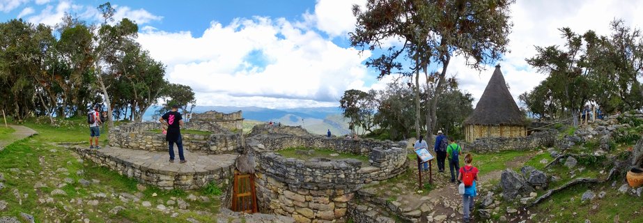 Panoramic view of Kuelap archaeological site showing ancient stone ruins, a thatched-roof structure, tourists walking on pathways, with mountains and cloudy sky in the background