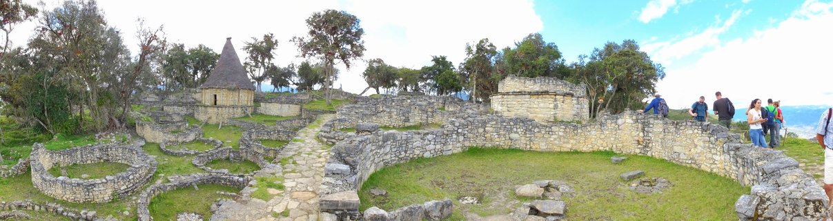 Panoramic view of Kuelap archaeological site showing ancient circular stone walls, reconstructed conical-roof buildings, stone pathways, green grass, trees, and tourists exploring the Chachapoya fortress ruins in Peru.