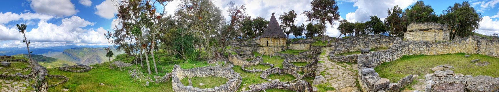 Ancient stone ruins of Kuelap fortress featuring circular foundations, stone pathways, and a conical-roofed building with green grass, trees, and mountains under a blue sky with white clouds