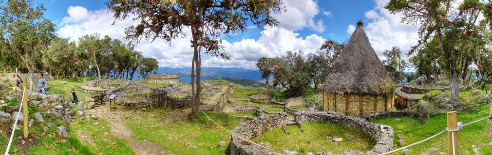 Panoramic view of ancient stone ruins at Kuelap archaeological site with circular walls, a thatched-roof reconstructed building, green grass, trees, and mountains under a blue sky with clouds