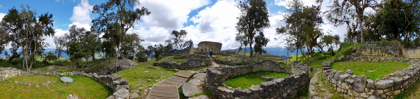Panoramic view of ancient circular stone wall ruins at Kuelap archaeological site in Peru, with grass-covered ground, wooden walkways, scattered trees, and mountains under a partly cloudy blue sky